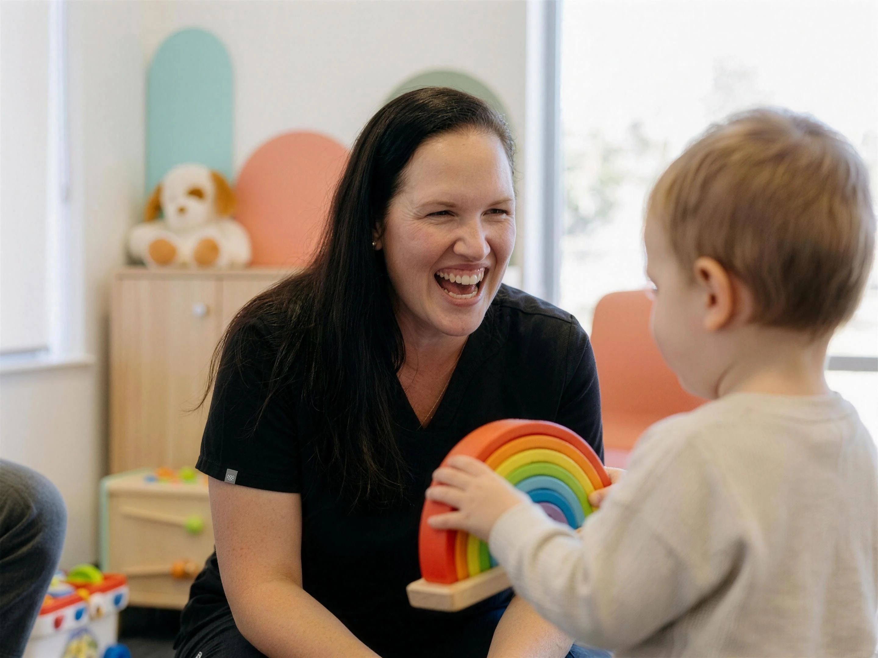 Dr O’Reilly in scrubs laughing with a toddler holding a rainbow toy
